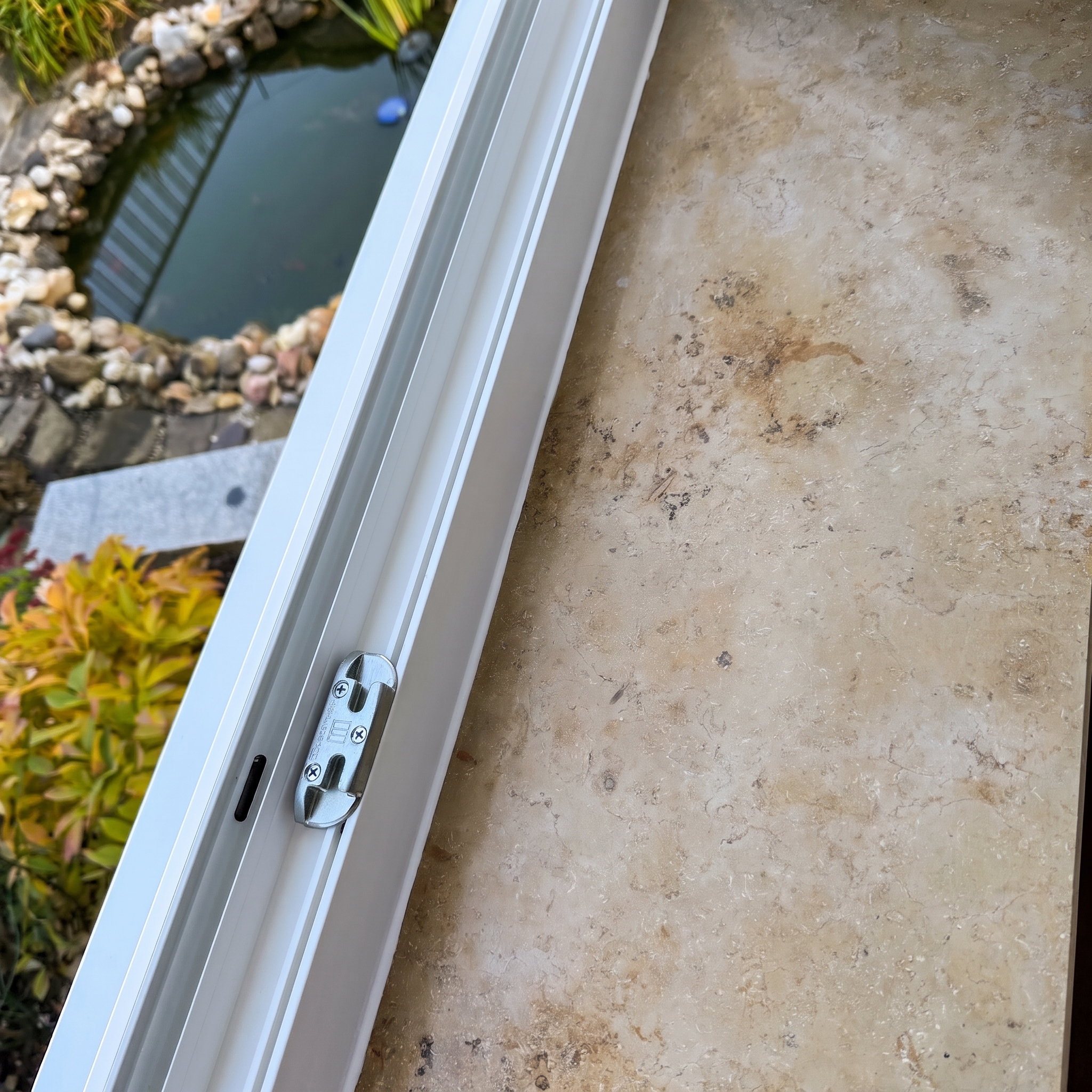 Close-up of a white sliding-window track with lock fitting, garden pond with stone edging behind